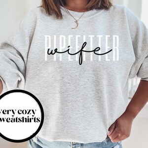 May include: Gray sweatshirt with the text "Pipefitter wife" in black lettering. The text is in a vertical format with white lines behind each letter.