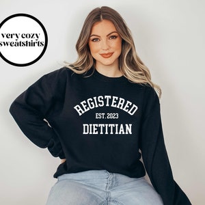 May include: Black sweatshirt with white text that reads "REGISTERED DIETITIAN EST. 2023". The sweatshirt also has a circular graphic that says "very cozy sweatshirts". The model is wearing blue jeans.