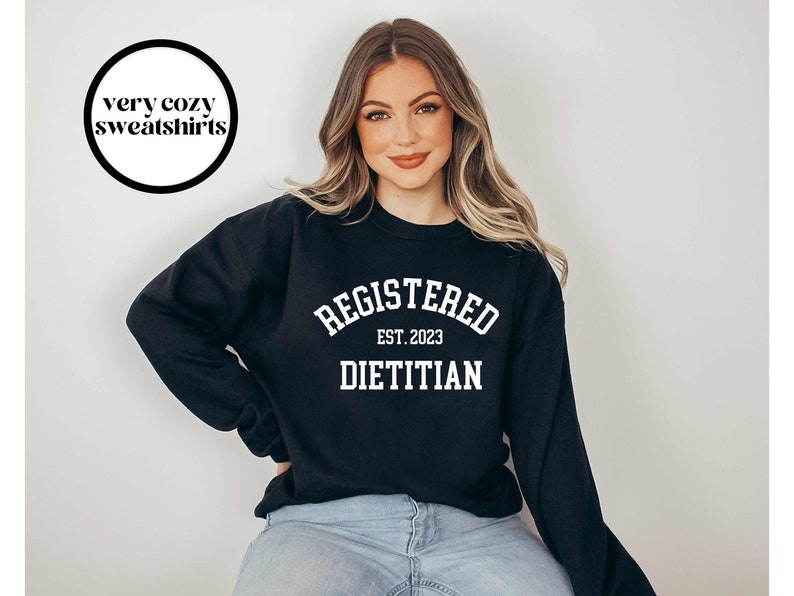 May include: Black sweatshirt with white text that reads "REGISTERED DIETITIAN EST. 2023". The sweatshirt also has a circular graphic that says "very cozy sweatshirts". The model is wearing blue jeans.