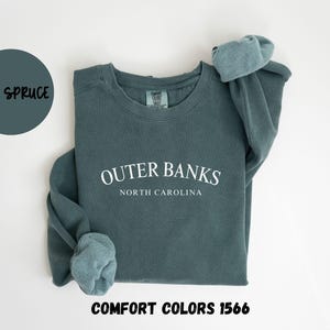 May include: A blue spruce-colored sweatshirt with the text "Outer Banks North Carolina" in white. The sweatshirt is a Comfort Colors 1566 style. A blue circle with the words "Blue Spruce" is in the upper left corner.