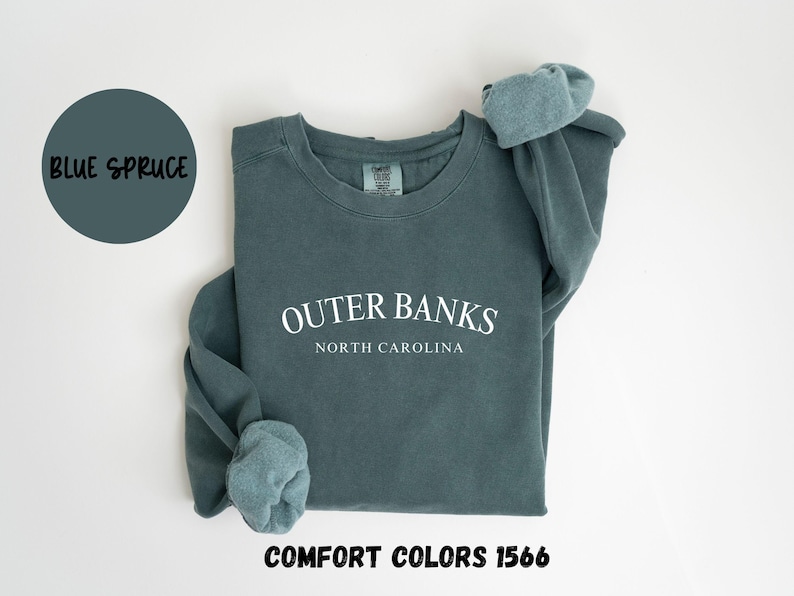 May include: A blue spruce-colored sweatshirt with the text "Outer Banks North Carolina" in white. The sweatshirt is a Comfort Colors 1566 style. A blue circle with the words "Blue Spruce" is in the upper left corner.