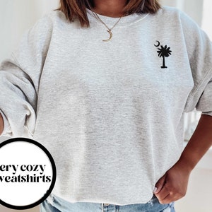 May include: A gray sweatshirt with a black palm tree and crescent moon design. The text "very cozy sweatshirts" is visible in a circle on the bottom left of the image.