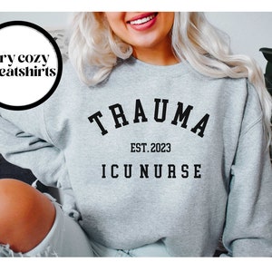 K&ouml;nnte beinhalten: Grauer Pullover mit schwarzem Text, der "Trauma Est. 2023 ICU Nurse" lautet.