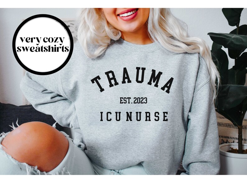 K&ouml;nnte beinhalten: Grauer Pullover mit schwarzem Text, der "Trauma Est. 2023 ICU Nurse" lautet.