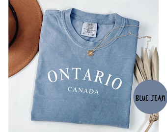 Puede incluir: Una camiseta azul vaquera con la palabra "ONTARIO" arqueada en la parte delantera, con "CANADA" debajo. Un collar dorado con un colgante descansa sobre la camisa. Un sombrero marrón es visible en la esquina superior izquierda.