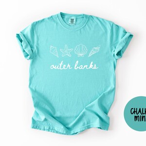 May include: A turquoise t-shirt with the words "outer banks" in white script, along with seashell and starfish graphics. The shirt has rolled-up sleeves and a round "Chalky Mint" logo.
