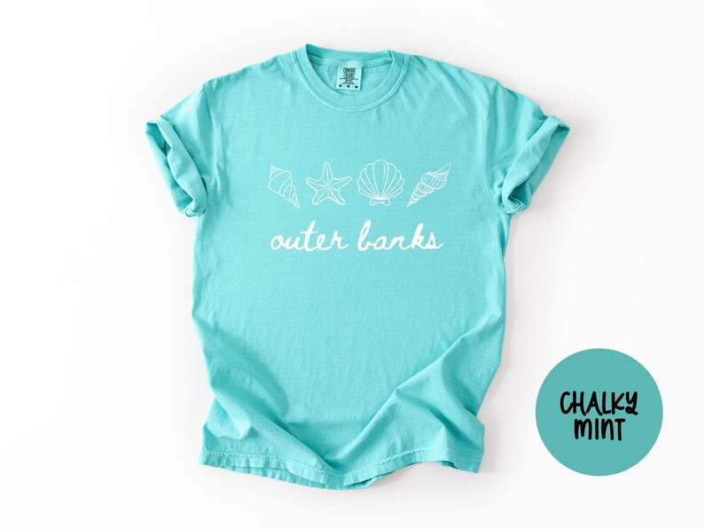 May include: A turquoise t-shirt with the words "outer banks" in white script, along with seashell and starfish graphics. The shirt has rolled-up sleeves and a round "Chalky Mint" logo.