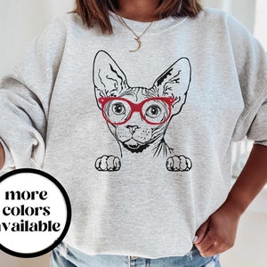 May include: Gray sweatshirt with a black and white illustration of a Sphynx cat wearing red glasses. The text "more colors available" is printed on a white circle in the lower left corner of the image.