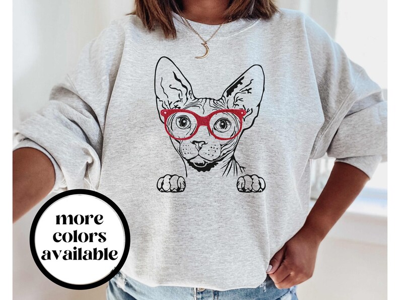 May include: Gray sweatshirt with a black and white illustration of a Sphynx cat wearing red glasses. The text "more colors available" is printed on a white circle in the lower left corner of the image.