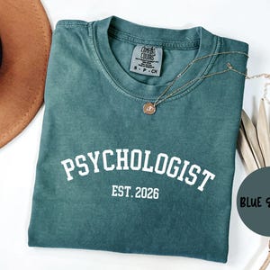 May include: Teal-coloured t-shirt with the word "PSYCHOLOGIST" in white block letters, with "EST. 2026" underneath. A gold necklace and a brown hat are also visible. The shirt is a Comfort Colors brand.