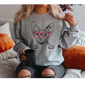 May include: A gray sweatshirt with a black and white illustration of a Sphynx cat wearing red glasses. The cat is peeking over the top of the sweatshirt.