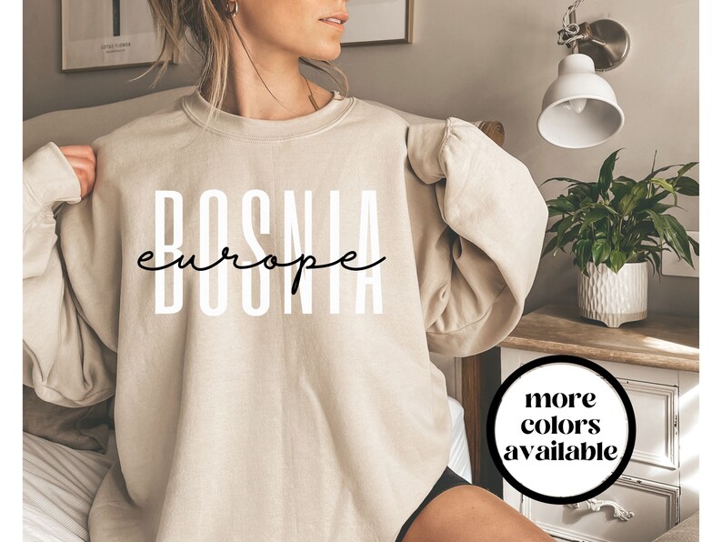 K&ouml;nnte beinhalten: Ein beiger Pullover mit dem Aufdruck "BOSNIA europe" in Schwarz auf der Vorderseite. Der Pullover wird von einer Person getragen. Der Text "mehr Farben verf&uuml;gbar" ist in Schwarz auf einem wei&szlig;en Kreis in der unteren rechten Ecke des Bildes gedruckt.