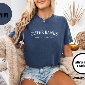 Puede incluir: Una camiseta azul marino con el texto "Outer Banks North Carolina" en blanco. La camiseta es parte de la colecci&oacute;n "Comfort Colors 1717" y se describe como "camisetas ultra suaves". Un c&iacute;rculo azul marino con la palabra "DENIM" est&aacute; en la esquina superior izquierda.