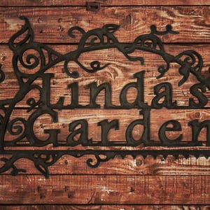 May include: Black metal sign with a decorative floral design and the text "Linda's Garden".
