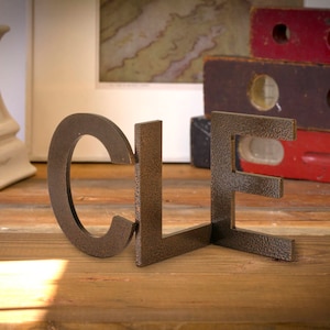 May include: Metal letters spelling out the word "CLE" in a rustic brown color. The letters are arranged in a staggered formation on a wooden surface.