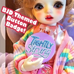 May include: A BJD doll holds a round, purple button badge with the words "Tightly Strung" in white script. The doll wears a colorful striped sweater in shades of pink, yellow, and blue. A pink accessory hangs from the badge.