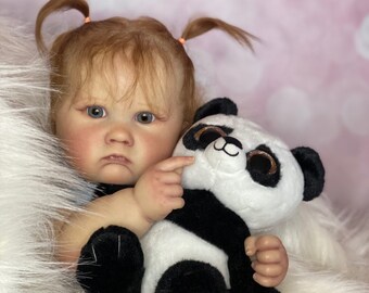 May include: A baby doll with red hair in pigtails holds a black and white panda plush toy. The doll has blue eyes and is wearing a white outfit. The doll and toy are sitting on a white fluffy surface.