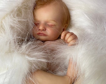 May include: A realistic baby doll with blonde hair and closed eyes, lying on a white fluffy blanket.