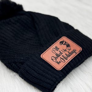 May include: Black knitted beanie with a faux fur pom pom and a brown leather patch that says "All Dolled Up for the Holidays".