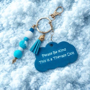 May include: A blue keychain with a gold clasp and ring. The keychain has a blue tassel and a cloud-shaped tag that reads "Please Be Kind This is a Therapy Doll." The keychain also features blue and white beads.