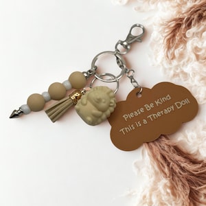 May include: A keychain with a brown tag that reads "Please Be Kind This is a Therapy Doll." The keychain includes a lion charm, a beaded accent, and a tassel. The metal hardware is silver.