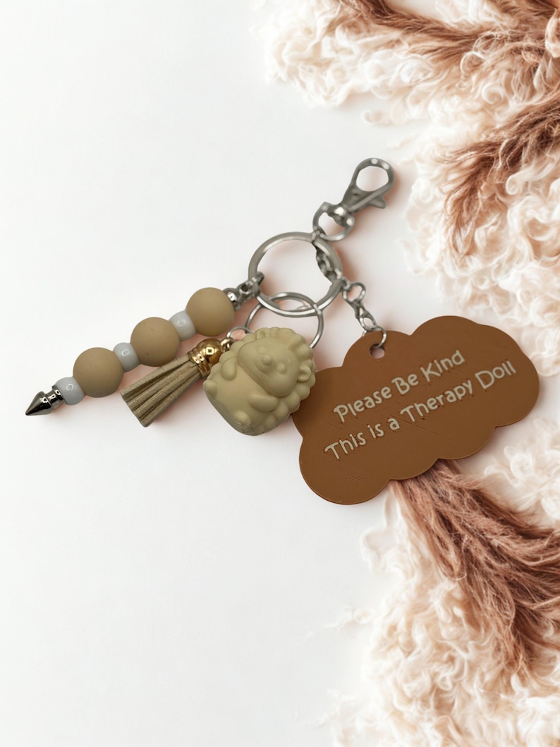 May include: A keychain with a brown tag that reads "Please Be Kind This is a Therapy Doll." The keychain includes a lion charm, a beaded accent, and a tassel. The metal hardware is silver.