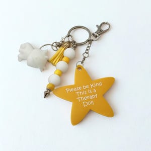 May include: A keychain with a yellow star-shaped charm that reads "Please be Kind This is a Therapy Doll." The keychain also features a white figurine, yellow tassel, white and yellow beads, and a silver clasp.