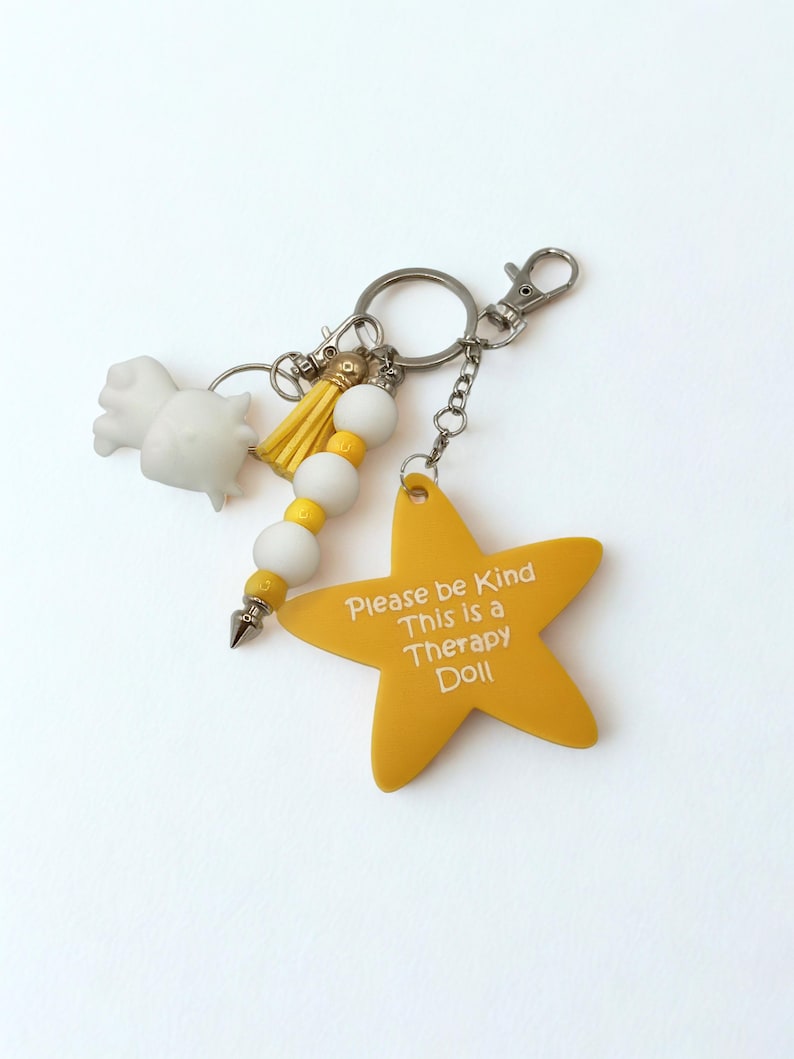 May include: A keychain with a yellow star-shaped charm that reads "Please be Kind This is a Therapy Doll." The keychain also features a white figurine, yellow tassel, white and yellow beads, and a silver clasp.