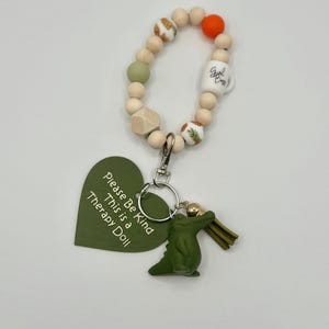 May include: A beaded keychain with a green alligator charm and a green heart-shaped tag that reads "Please Be Kind This is a Therapy Doll." The keychain includes wooden beads, a white mug bead, and an orange bead.