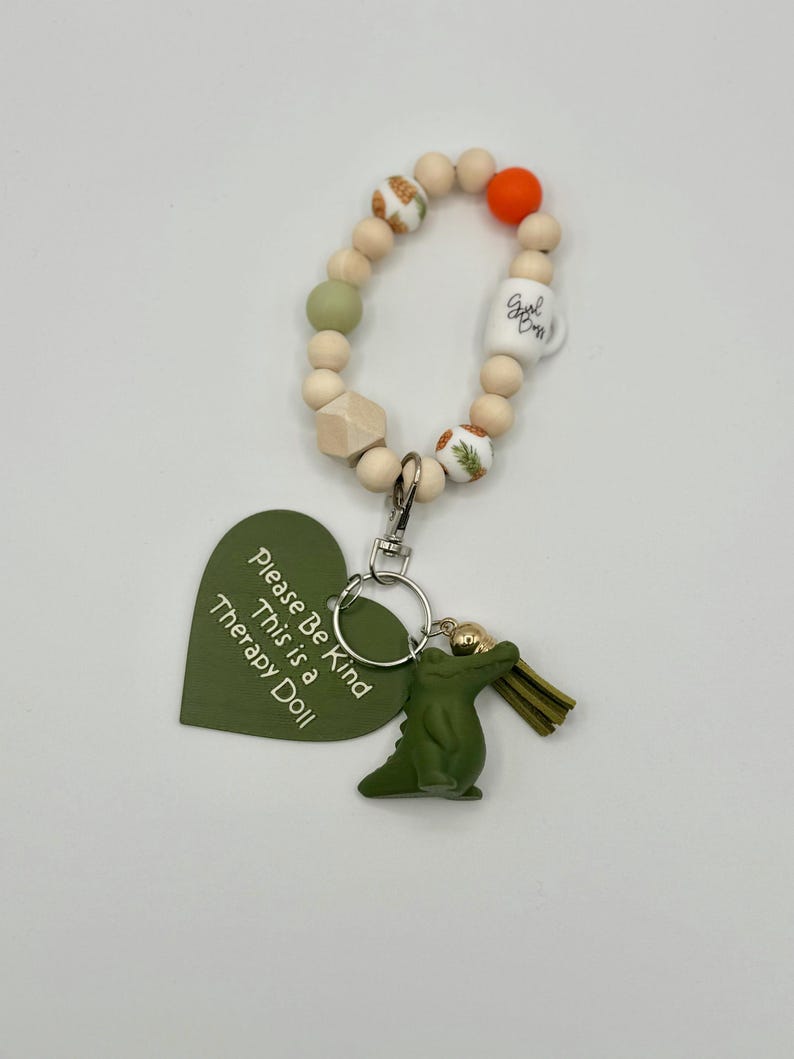 May include: A beaded keychain with a green alligator charm and a green heart-shaped tag that reads "Please Be Kind This is a Therapy Doll." The keychain includes wooden beads, a white mug bead, and an orange bead.
