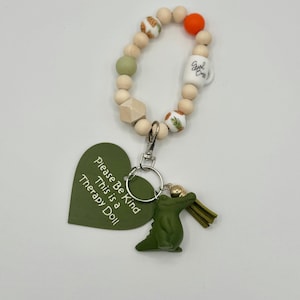 May include: A beaded keychain with a green alligator charm and a green heart-shaped tag that reads "Please Be Kind This is a Therapy Doll." The keychain includes wooden beads, a white mug bead, and an orange bead.