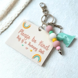 May include: A keychain with a light pink tag that reads "Please be kind This is a Therapy Doll" with rainbow and heart accents. The keychain has pink, white, yellow, and green beads, a silver-colored charm, and a light blue dinosaur charm.
