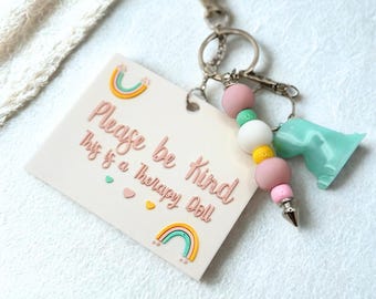 May include: A keychain with a light pink tag that reads "Please be kind This is a Therapy Doll" with rainbow and heart accents. The keychain has pink, white, yellow, and green beads, a silver-colored charm, and a light blue dinosaur charm.