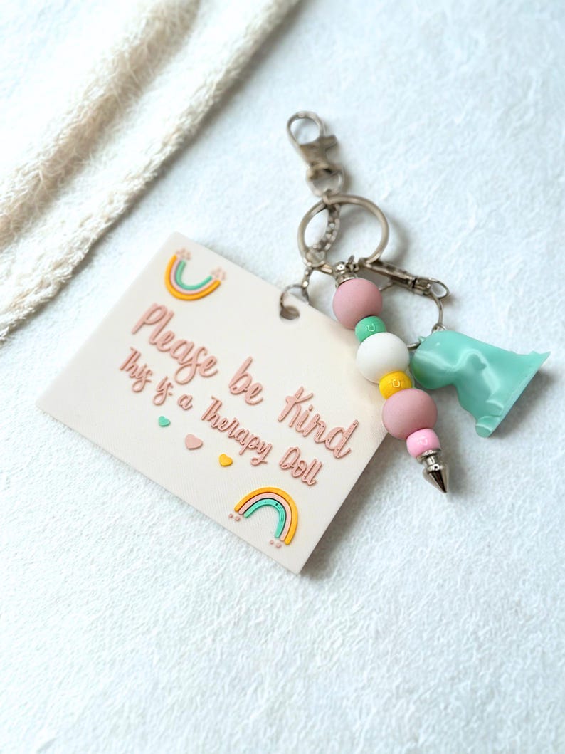 May include: A keychain with a light pink tag that reads "Please be kind This is a Therapy Doll" with rainbow and heart accents. The keychain has pink, white, yellow, and green beads, a silver-colored charm, and a light blue dinosaur charm.