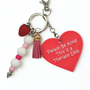 May include: A keychain with a red heart-shaped charm that reads "Please Be Kind This is a Therapy Doll." The keychain also features a red strawberry charm, a pink tassel, and a beaded accent with white and pink beads.