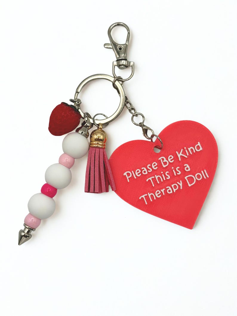 May include: A keychain with a red heart-shaped charm that reads "Please Be Kind This is a Therapy Doll." The keychain also features a red strawberry charm, a pink tassel, and a beaded accent with white and pink beads.