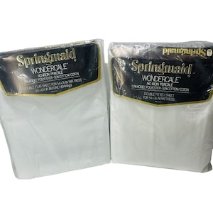 Könnte beinhalten: Zwei versiegelte Packungen Springmaid Wondercale Bettlaken. Die Verpackung hat ein schwarz-goldenes Etikett mit dem Markennamen und Produktdetails. Die Laken sind weiß und für eine Matratze von 137x193 cm konzipiert.