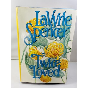 Puede incluir: Un libro titulado "Twice Loved" de LaVyrle Spencer. La portada presenta crisantemos amarillos y hojas verdes sobre un fondo crema. El título y el nombre del autor son de color azul.