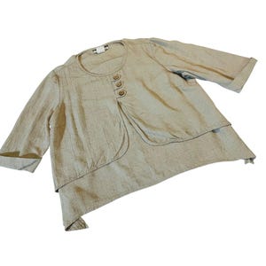 Puede incluir: Top beige de mezcla de lino con un diseño en capas. La parte superior presenta un cuello redondo, tres botones y mangas tres cuartos. El dobladillo asimétrico añade un toque único a esta prenda casual.