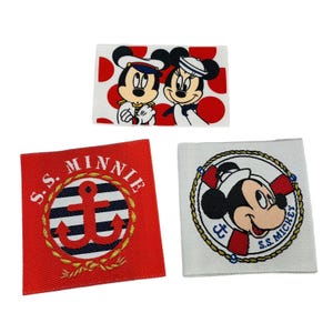 Puede incluir: Tres parches de tela roja y blanca con imágenes bordadas de Mickey Mouse y Minnie Mouse. El primer parche presenta a Mickey y Minnie con atuendos de marinero. El segundo parche presenta un ancla roja con un fondo rayado blanco y negro. El tercer parche presenta a Mickey Mouse con un atuendo de marinero con el texto "S.S. MICKEY".