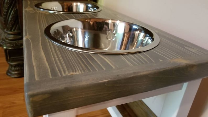 K&ouml;nnte beinhalten: Ein grauer Holz-Hunde-Napfst&auml;nder mit zwei Sch&uuml;sseln aus Edelstahl. Der St&auml;nder hat eine wei&szlig;e Basis und ist aus recyceltem Holz gefertigt.