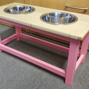 K&ouml;nnte beinhalten: Ein erh&ouml;hter Hundenapfst&auml;nder aus rosa Holz mit zwei Sch&uuml;sseln aus Edelstahl. Der St&auml;nder hat eine Ablage darunter f&uuml;r die Aufbewahrung.