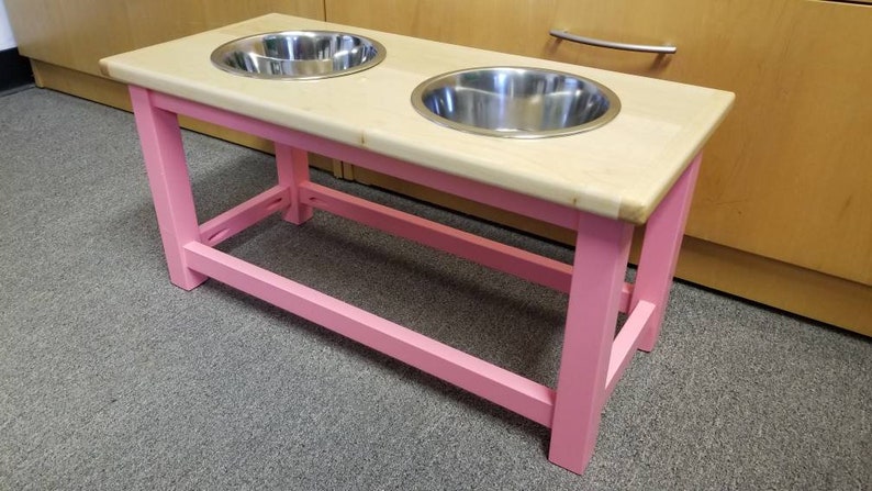 K&ouml;nnte beinhalten: Ein erh&ouml;hter Hundenapfst&auml;nder aus rosa Holz mit zwei Sch&uuml;sseln aus Edelstahl. Der St&auml;nder hat eine Ablage darunter f&uuml;r die Aufbewahrung.