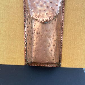 May include: A handcrafted copper wall pocket with a textured surface and a flap closure. The rectangular pocket is riveted along the edges and has a dark cord for hanging. The copper has a warm, reddish-brown hue.