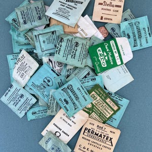 May include: A collection of small, vintage watch crystal packets in various colors, including teal, beige, and white. The packets feature printed text and logos, with some labeled "GS Flexo" and "Permatex". The items are scattered on a blue surface.