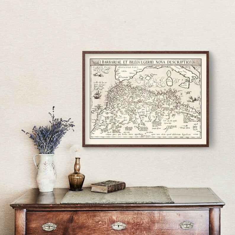 K&ouml;nnte beinhalten: Eine Vintage-Karte der Barbarenk&uuml;ste und der umliegenden Gebiete, gerahmt in einem braunen Holzrahmen. Die Karte ist in Schwarzwei&szlig; gedruckt und zeigt eine Vielzahl von geografischen Merkmalen, darunter St&auml;dte, Fl&uuml;sse und Berge. Der Titel der Karte lautet "I BARBARIAE ET BILEDVLGERID NOVA DESCRIPTIO".
