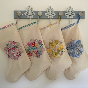 May include: Four cream Christmas stockings with floral patchwork designs. Each stocking features a different floral pattern in pinks, blues, yellows, and reds. They hang from a grey wall-mounted rack with decorative hooks.