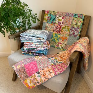 May include: A stack of colourful patchwork quilts in various floral patterns, including pink, blue, green and yellow, are displayed on a wooden chair with a light grey cushion.