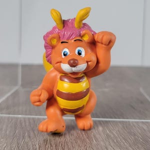 Könnte beinhalten: Kleine, orangefarbene Figur einer Zeichentrickfigur mit Löwenmähne und bienenartigen Streifen auf dem Bauch. Die Figur hat blaue Augen, gelbe Hörner und hebt eine Hand. Die Figur steht auf einer grauen Oberfläche.