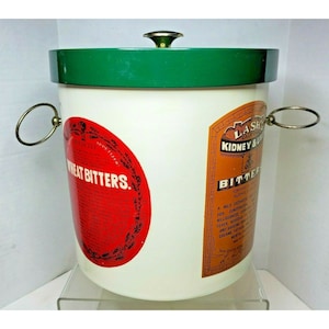 May include: A white metal ice bucket with a green lid and two metal handles. The bucket has two labels, one for "Wheat Bitters" and one for "Lash's California Kidney & Liver Bitters".